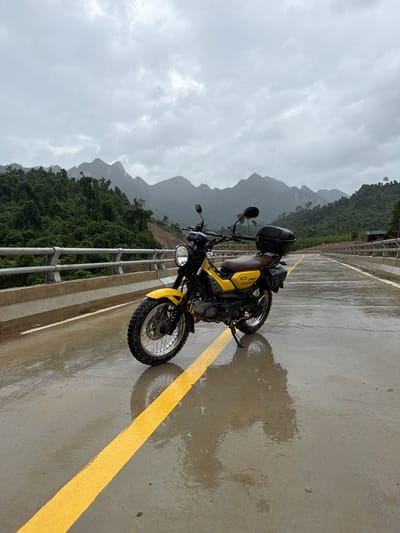 A yellow Yamaha motorcycle is parked on a wet, paved road in Trường Sơn Commune, Vietnam, under a cloudy sky. The road has a yellow dividing line and reflections of the motorcycle and surroundings are visible on the wet surface. The motorcycle is positioned slightly to the left of the frame, facing towards the right. It has a black storage box on the rear and saddlebags on the sides. The background features lush green, forested mountains, a concrete bridge railing, and a clear sky with scattered clouds. The overall scene depicts a rural, possibly remote, location during overcast weather.