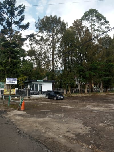 The image shows an outdoor scene with various natural and man-made elements. A parking lot with a dark-colored car parked near a small, white building labeled "POS JAGA" (Guard Post) is visible. Behind the building, there are several trees, and a fence runs along the background. A sign indicates the location as "PARKIR WISATA ALAM AIR TERJUN CIBEUREUM" (Cibeureum Waterfall Natural Tourism Parking Area), part of the "TAMAN NASIONAL GUNUNG GEDE PANGRANGO" (Gunung Gede Pangrango National Park). An orange traffic cone is placed near the sign, and the foreground shows a damaged asphalt road. The environment appears to be a mountainous area, as suggested by the trees and the sign mentioning Gunung Gede Pangrango. The image doesn't indicate any people or interactions. The location in the city of Mya Sit Kaing Ywar Thit, Myanmar, is not verifiable from the image alone and seems unrelated to the provided context of the image.