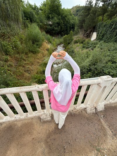 A person wearing a white hijab, a pink long-sleeved shirt, and white pants stands on a bridge, forming a heart shape with their hands above their head. The bridge has a white, weathered railing with visible metal pipes underneath. Below the bridge, a stream flows through a lush, green valley with trees and vegetation covering the slopes of a hill. The scene appears to be in the city of Khumsan, Uzbekistan.