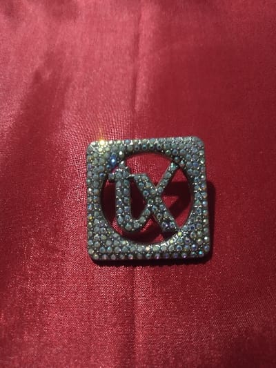 The image depicts a close-up, top-down view of a rectangular decorative object, approximately 4-5 cm in dimension. This object, likely a brooch or buckle, features a stylized 'TX' monogram cutout within a square frame with slightly rounded corners. Both the frame and the letters are densely encrusted with numerous small, faceted, clear or iridescent rhinestones, creating significant sparkle and exhibiting multi-colored light reflections. The underlying material of the object, partially visible between the crystals, appears silver-toned. The object rests on a deep crimson red fabric surface, which shows visible folds and a smooth texture consistent with satin or silk. No individuals, actions, or additional specific objects are present in the immediate scene. The location of this scene is Horažďovice, Czechia.