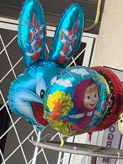 A cluster of three foil balloons is suspended outdoors, likely outdoors, against a wall and a window. The balloons are a Bugs Bunny-shaped balloon with large ears and a face on the main body, a round balloon with an image of the character Masha from the animated series "Masha and the Bear" on a blue background with trees and clouds, and a partially visible red balloon with a black and white patterned edge. The Bugs Bunny balloon is blue and features images of Bugs Bunny holding a carrot on each ear. The Masha balloon has Masha in a pink headscarf and dress. The balloons appear to be tied together with white string and a pink ribbon. A metal railing or shelf is visible below the balloons. The background consists of a textured wall, a window with a grid, and a yellow pipe. The scene suggests a festive or celebratory occasion. The location is Orhei, Moldova.