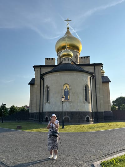 A woman stands on a cobblestone path in front of a large building with golden domes. She is wearing a white hat, a patterned dress, and white shoes, and is holding a cane and a phone. She appears to be taking a picture of herself or the building. The building, a church, has light-colored stone walls and a dark roof. The sky is blue with some wispy white clouds, suggesting it is daytime. The scene is well-lit, indicating it's likely sunny. The ground is a mix of cobblestones and green grass. The overall atmosphere is peaceful and serene. There is no visible text in the image. The location is identified as Kirishi, Russia.