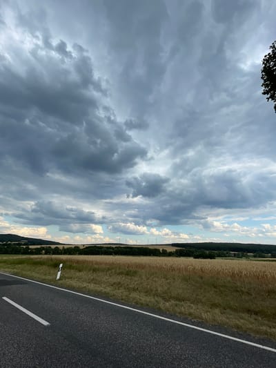 The image shows a rural roadside scene in Holzburg, Germany. A dark asphalt road with a white line marking runs diagonally from the bottom right towards the center of the frame. To the left of the road, a field of dry, golden grass stretches into the distance, bordered by a dense line of dark green trees. In the far distance, on the horizon, several wind turbines are visible, suggesting an open, agricultural landscape. Above the field and trees, the sky is filled with dramatic, textured clouds. The clouds are a mix of dark grays and lighter whites, indicating a dynamic and possibly stormy weather pattern. The light suggests it is late afternoon or early evening. There are no people or specific objects of note in the scene, aside from the road markings and wind turbines. The overall impression is one of a natural, expansive landscape under a moody sky.