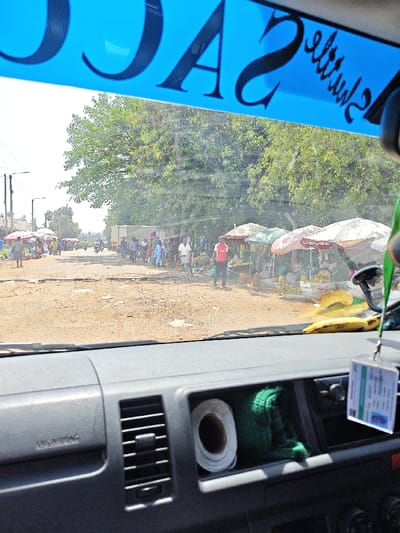 The interior of a car shows a dashboard with a roll of paper and a green object in a compartment. Through the windshield, a busy street market scene unfolds. People stroll along a dusty road lined with vendors under umbrellas, selling fruits and vegetables. A white truck is parked in the distance. A blue sign with large letters is partially visible across the top of the frame.