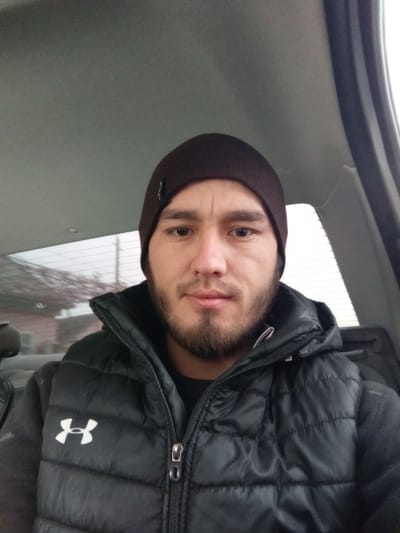 A man sits inside a car wearing a brown beanie and black Under Armour vest. He has short brown hair and a thick beard. The car's interior is visible, including the ceiling, window, and back of a seat. The man looks directly at the camera with a neutral expression. The light suggests it could be daytime, potentially overcast.