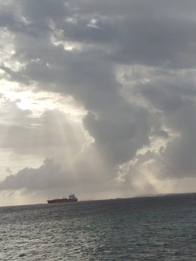 A wide shot captures the ocean and sky on a cloudy day in Gulhi Falhu, Maldives. The sea, with its rippling surface, occupies the lower half of the frame. On the horizon, a large, orange cargo ship is visible, sailing through the water. Above the sea, the sky is filled with dramatic, grey clouds, with shafts of sunlight breaking through in some areas, creating a sense of an approaching storm or post-storm glow. In the distance, along the horizon, faint outlines of buildings suggest the presence of land.