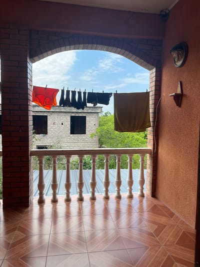 The image shows a balcony with a brick archway opening to an outdoor scene. Laundry, including an orange garment and several dark items, hangs on a clothesline strung across the opening. Beyond the balcony, a partially constructed building with dark window openings is visible, surrounded by green trees. The balcony floor is tiled with a brown geometric pattern, and a white balustrade runs along the edge. The right wall of the balcony is textured and orange, with a small wooden shelf and a decorative circular object mounted on it. The sky above is blue with scattered white clouds. The scene appears to be a residential area in the municipality of Ghvranekeli, Georgia.