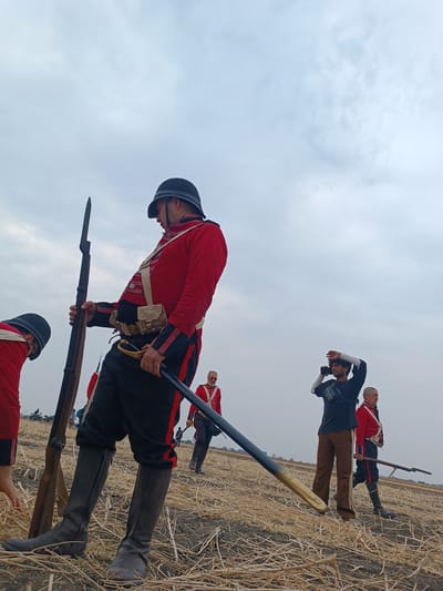 The scene depicts an open field in Kottalapalli, India, under an overcast sky. In the foreground, a male figure dressed in a red military-style coat with black trim, black trousers with red stripes, a black helmet, and tall dark boots stands, holding a long rifle with an attached bayonet upright in his left hand and a scabbarded saber with a brass hilt in his right. He wears a beige canvas belt with a pouch. To his immediate left, another individual in similar attire is partially visible, bending over.
In the midground, a male individual in contemporary clothing, consisting of a black long-sleeved shirt, brown pants, white arm sleeves, and a black cross-body bag, holds a black smartphone horizontally near his head, appearing to film or observe. To his right, another male figure in a red military-style coat, black trousers, and boots walks towards the right, holding a rifle. Further in the background, a man with a white beard and glasses, also in a red uniform, is partially visible.
The ground is a dry, harvested field covered with straw stubble and patches of exposed dark soil. Distant, indistinct figures and what appear to be motorcycles are visible along a far horizon line. The sky is uniformly light grey with cloud cover.