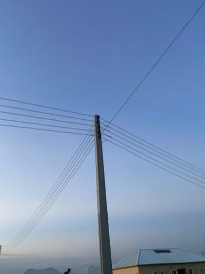 The image shows a low-angle view of power lines against a clear sky. A concrete utility pole with multiple wires extending from it is visible in the foreground, running diagonally across the frame. In the background, a light-colored building with dark window frames is partially visible. The sky is a gradient of light blue, suggesting daylight, possibly late afternoon or early morning. There is no visible text in the image. The scene appears to be an outdoor setting, likely in an urban or semi-urban environment given the presence of the utility pole and building. There are no people or animals depicted. The overall impression is one of utility infrastructure against a calm, clear sky.