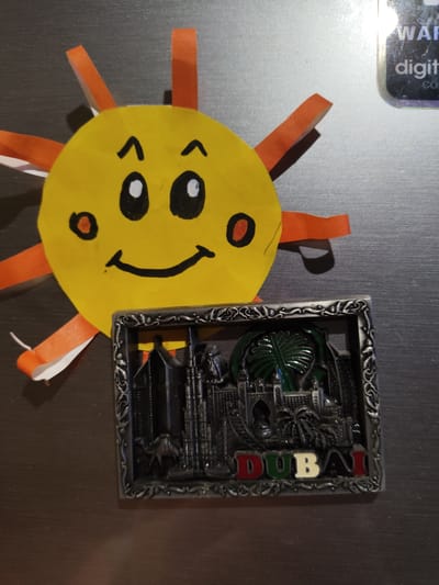 A sun drawing and a Dubai souvenir magnet are affixed to a surface. The sun drawing is yellow with orange rays, and a smiling face drawn on it. The Dubai magnet is square with an ornate frame. "DUBAI" is spelled out across the bottom with each letter in a different color. Buildings and a palm tree are molded in relief above the lettering. The background surface appears to be a dark laminate. The location is not specified in the city of Yerzhovo, Moldova.