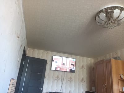 The image shows a residential interior, likely a living room in Kuznetsk, Russia. The scene features a dark grey door on the left side of the frame, with a thin white vertical stripe. To the right of the door, a television is mounted on a wall covered in patterned wallpaper. The TV displays a scene from what appears to be a courtroom drama, with people seated at desks and a central figure at a podium.
In the upper right portion of the image, a ceiling light fixture with a classic, ornate design is visible, featuring clear crystal accents and a white lampshade. Further to the right, a tall wooden cabinet or wardrobe stands against the wall, with two doors that have curved detailing. The wallpaper in this area also has a repeating pattern, featuring floral or scroll motifs.
The walls appear to be a light color, with some areas showing signs of wear or imperfections, particularly on the left side of the frame where the plaster seems to be exposed in places. The overall lighting suggests it is daytime, though the specific time of day is not evident from the image. There are no visible signs of weather conditions. The room appears to be a private residence. No specific emotions are conveyed by the scene. There is no visible text in the image.