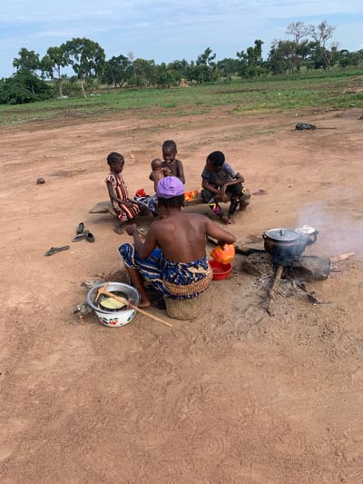 In Nandu, Nigeria, a group of individuals gathers around an outdoor cooking fire. A woman, wearing a blue and white patterned skirt and a purple headwrap, is seated on a log, tending to a cooking pot over the fire. She holds an orange bowl in her right hand, and a red bowl is near her left. To her left, a metal bowl with a cherry pattern contains a yellow liquid and a wooden utensil. Two young children are seated on a wooden plank to the left of the woman. One child, dressed in a striped dress, is looking down, while the other, holding a baby, is looking towards the camera and smiling. A third child, wearing a striped shirt and pants, sits further down the plank, looking to the right. Several pairs of shoes are scattered on the ground. In the background, a field with greenery and trees stretches towards the horizon under a cloudy sky. A black object, possibly a rolled-up mat or bag, lies on the ground in the middle distance. The ground is predominantly red dirt.