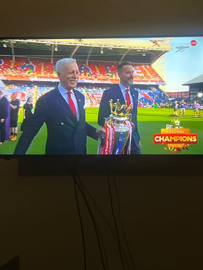 The image shows a television screen displaying a scene of Arsenal celebrating a Premier League championship, with a "Champions 2025/26" graphic. Two men in suits are holding the Premier League trophy. The background shows a stadium filled with fans. The Premier League logo is visible on the screen. The location is not Oshogbo, Nigeria. The scene is from a Premier League stadium during a championship celebration.