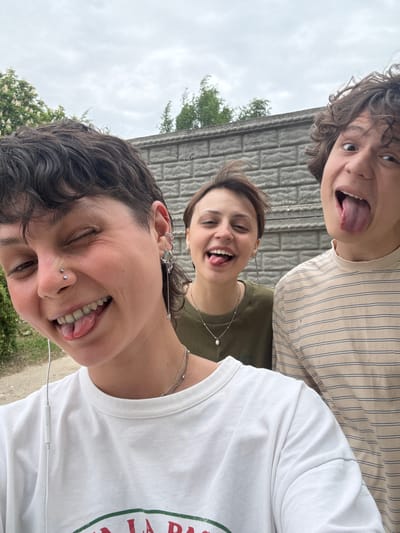 Three people are posing for a selfie. The photo is taken from a low angle, looking up at the subjects. The person in the foreground is wearing a white t-shirt and has dark curly hair. They have a nose piercing and multiple ear piercings. They are sticking their tongue out. The person in the middle is wearing a green shirt and a silver necklace. They are also sticking their tongue out. The person in the background is wearing a tan and white striped shirt and has curly brown hair. They are sticking their tongue out.
In the background, there is a concrete wall. There are some trees and greenery visible. The sky is overcast. It is impossible to verify if the location is Uryupinsk, Russia.