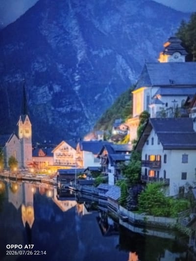 The image depicts a twilight scene of a picturesque town nestled at the base of a large, dark blue mountain. The mountain dominates the upper portion of the frame, with its rugged texture visible in the dim light. In the middle ground, a cluster of traditional European-style buildings, with steep roofs and illuminated windows, line the waterfront. A prominent church with a tall steeple stands out on the left side of the scene, its structure illuminated by warm light. The buildings reflect in the calm, dark water of a river or lake that occupies the lower half of the image. The overall impression is one of serene beauty and architectural charm. The bottom left corner of the image displays text indicating "OPPO A17" and a date and time stamp: "2026/07/26 22:14". There is no indication of the city of Құмөзек, Kazakhstan in the image.