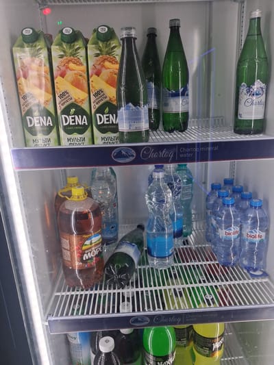 This is a photograph taken inside a refrigerator or cooler displaying a variety of beverages. The subjects are primarily bottles and cartons of drinks, arranged on shelves.
The drinks visible include several green glass bottles with white labels that have blue accents and imagery of mountains and flowers, suggesting they contain mineral water or a similar beverage. There are also clear plastic bottles with blue caps, some labeled "biolife active." Additionally, there are cartons of juice, prominently featuring the brand "DENA" and text that appears to be in Cyrillic, indicating "100% NATURAL FRUIT" and "MULTI FRUIT." Another large plastic bottle is visible with a yellow cap and a label with Cyrillic text, suggesting a juice or beverage with a dark liquid.
The setting is the interior of a commercial refrigerator, characterized by white slatted shelves and clear glass doors. The lighting suggests an indoor environment, likely a store or supermarket, possibly in a location like Kahramon, Uzbekistan, based on the Cyrillic text on the products. There is no discernible time of day or weather information. The overall mood is neutral, focusing on the products being offered. The prominent text includes brand names like "Chortog," "biolife," "DENA," and "MOPO," as well as descriptions of the contents such as "mineral water" and "multi fruit."