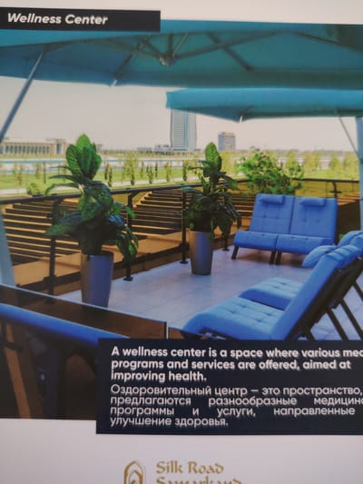 The image displays a modern outdoor seating area, presumably part of a wellness center. In the foreground, two blue loungers and a blue sofa are arranged on a tiled patio. Two large potted plants with lush green leaves flank the seating area. In the background, tiered steps lead down to a manicured landscape with trees and a body of water, possibly a river or lake. Beyond the greenery, modern buildings and skyscrapers are visible, suggesting an urban setting. A dark banner across the top of the image reads "Wellness Center". Below the seating area, a dark rectangular panel contains text in both English and Cyrillic. The English text reads: "A wellness center is a space where various medical programs and services are offered, aimed at improving health." The Cyrillic text is partially visible and appears to be a translation of the English text. The bottom of the image shows a logo that reads "Silk Road". The overall impression is one of a serene and upscale facility designed for relaxation and health improvement. The mention of "Bahshitpa, Uzbekistan" is not directly evident from the visual cues in the image itself.