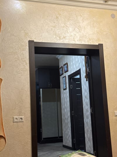 The image shows an interior view through a doorway. The doorway has a dark brown frame and leads into a hallway with a textured wallpaper on the walls. Inside the hallway, there is a closet with mirrored sliding doors, and a dark brown door that is slightly ajar. On the wall to the left of the doorway, there is a white electrical outlet with two sockets. The wall in the foreground, to the left of the doorway, has a beige textured wallpaper with an abstract pattern. The lighting suggests it is daytime, with ambient light entering from the hallway. The scene appears to be a residential interior. There are no people visible in the image. The location in Ptghni, Armenia cannot be determined from the visual information in the image.