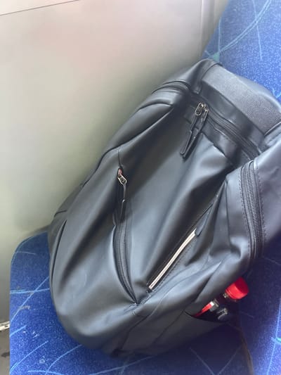 A black backpack rests on a blue, patterned seat, possibly in a public transport setting. A red-capped bottle is partially visible in an outer pocket of the backpack. The background consists of a light-colored wall. The image does not provide any information about the city of Kashgar mahalla, Uzbekistan.