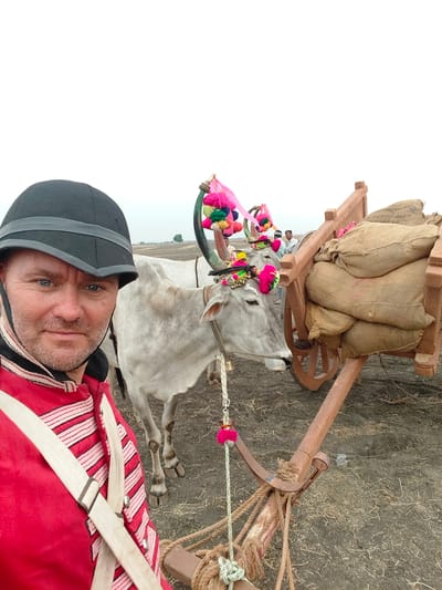 A man in a black helmet and a red jacket with white striped details and a white cross-belt takes a selfie in Kottalapalli, India. He has blue eyes and stubble. To his right and behind him are two light gray or white oxen, each adorned with colorful tassels and small bells on their horns and foreheads. The oxen are yoked to a rustic wooden bullock cart, which is loaded with several large, light brown burlap sacks. The cart has wooden wheels and a simple, sturdy construction. In the mid-ground, beyond the cart, several other men are partially visible, observing the scene. The ground is dry and dusty, covered with sparse, light-colored dry grass or stubble. The sky is overcast and light, indicating an outdoor, possibly rural or semi-rural setting. A few distant trees are visible on the horizon under the bright, cloudy sky.