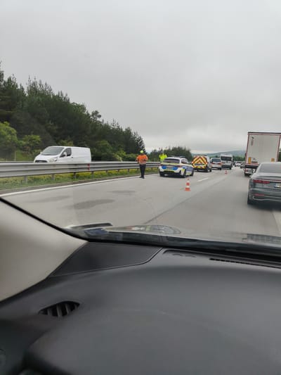 A traffic incident is unfolding on a highway, with emergency services present. A police car, an ambulance, and what appears to be a van are stopped on the right side of the road. Two individuals in high-visibility vests are standing near the police car, and another person in similar attire is positioned further back, nearer to the oncoming lane. Traffic cones have been placed on the road to redirect vehicles. The highway is bordered by a dense line of trees on the left and extends into the distance under an overcast sky. The foreground of the image is dominated by the dashboard and windshield of a vehicle, suggesting the photo was taken from a car. Based on the presence of emergency vehicles and the traffic cones, this scene is consistent with an accident or a police operation on a highway. The location is identified as Stolnik, Bulgaria.