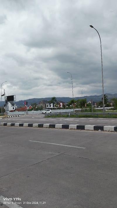 An outdoor, eye-level shot captures a road intersection or merge point in Sumedang, Indonesia, under an overcast sky. In the foreground, a wide asphalt road stretches across the bottom of the frame, featuring white road markings and a concrete median barrier with alternating black and white blocks.
In the midground, a white compact car is visible on a road that appears to curve to the left, possibly at a junction. To the left, a traffic light pole with a black rectangular structure, possibly a billboard or digital sign, stands next to a white building. Several streetlights with tall, curved poles are distributed throughout the scene, reaching towards the sky. A metal guardrail runs along the right side of the road, separating it from an elevated area.
The background reveals a residential area with multiple houses, some featuring red roofs and others predominantly white, built on gentle slopes. Behind the houses, green, undulating hills extend into the distance, partially obscured by the heavy cloud cover. The sky is uniformly grey and cloudy, indicating an overcast day.
A watermark in the bottom left corner displays "vivo Y19s", "Akhtar & Arvan", and the date and time "31 Des 2024, 11.29". The scene appears quiet with minimal traffic.