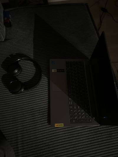 The image depicts a dark, dimly lit room, possibly a bedroom or living area, with a ribbed, dark gray textured surface that appears to be a sofa or bed. On this surface, a pair of black headphones is placed to the left. To the right of the headphones is a closed gray laptop with a yellow Lenovo sticker on its lower right corner. The laptop is positioned at an angle, with its screen slightly ajar, revealing a portion of the keyboard. A dark shadow, cast from an unseen light source, dominates the upper portion of the image. The presence of a power cord and a black adapter near the laptop suggests it is plugged in. The overall impression is one of a quiet, personal space. There is no indication of the city of Siedlce, Poland, or any specific location within it.