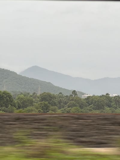 The image depicts a hazy landscape dominated by rolling, green hills in the background. The sky is overcast, creating a diffused light. In the mid-ground, a dense forest of trees, including palm-like varieties, covers the lower slopes of the hills. A few tall electrical pylons are visible within the treeline, suggesting the presence of infrastructure. On the far right, partially obscured by trees, are modern-looking buildings with white facades, indicating a developed area or settlement within the natural setting. The foreground is blurred due to motion, showing what appears to be a railway track and the ground beside it. This motion blur suggests the photograph was taken from a moving vehicle, likely a train, traveling along a route that passes through this scenic, hilly terrain near Vasai-Virar, India.