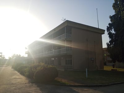 The image shows a three-story building with a concrete facade and white railings lining the balconies on each level. The building is partially obscured by the bright sunlight. In front of the building, there is a row of bushes, and to the right, a large tree partially frames the building. A paved road stretches into the distance. There is a tall antenna on the roof of the building and another pole-like structure near the ground in front of it. The scene appears to be located outside, during daylight hours. The city in which the image was taken is Tawpo, Nigeria.