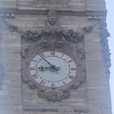 The image shows a close-up of an ornate clock face on the exterior of a stone building. The clock features Roman numerals, with the number XII at the top and VI at the bottom. The clock hands are visible, with the hour hand pointing to approximately X and the minute hand pointing to approximately IV. Above the clock, there is a sculpted bust of a man wearing a papal tiara and ornate robes. Decorative elements of foliage and carved stone surround the clock and the bust. Below the clock, an inscription in stone reads "LEONE XIII". The building appears to be constructed of light-colored stone blocks. The overall scene is of an architectural detail, likely a monument or a historical building, in Calcinate, Italy, identified by the inscription. The sky is visible in the upper right, appearing overcast.