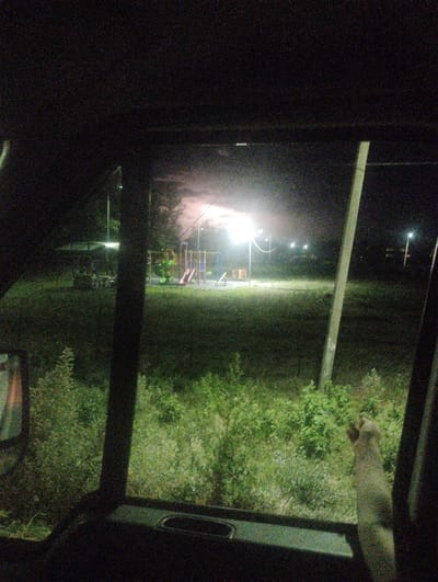 The image captures a nighttime scene viewed from inside a vehicle, looking out through the windshield and a side window. In the distance, a playground is illuminated by bright lights, with swings and slides visible. A covered picnic area is situated to the left of the playground. The surrounding area is covered in grass and dense green foliage, with several utility poles dotting the landscape. The sky above is dark but illuminated by the bright lights from the playground and possibly distant streetlights, creating a hazy glow. The foreground shows parts of the vehicle's interior, including a dark dashboard and a portion of a seat. A light-colored arm or limb, possibly a dog's, is visible in the right side of the frame, reaching out or resting near the window. The overall atmosphere suggests a rural or park setting at night, possibly in Akhali Dioknisi, Georgia, given the location context. There is no visible text in the image.