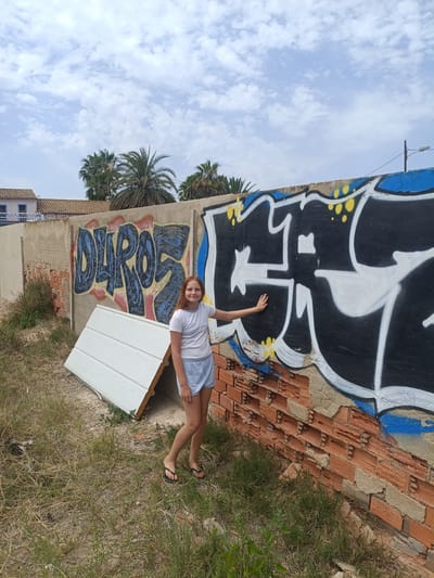 A young girl with auburn hair and fair skin stands in front of a graffiti-covered brick wall in Almardà, Spain. She is wearing a white t-shirt and light blue shorts, with black flip-flops on her feet. Her left arm is extended and her hand is resting on the graffiti. The graffiti on the wall includes the word "DUROS" in large blue letters with pink and yellow accents, and a larger black and white design to the right of it. Part of the wall is crumbling brick, while other sections are concrete.
To the left of the girl, a large white object, possibly a piece of a fence or a panel, is leaning against the wall. In the background, palm trees and buildings are visible under a cloudy sky. The scene appears to be outdoors during the daytime, with dry grass and weeds covering the ground. The girl is smiling at the camera, conveying a relaxed and positive emotion.
