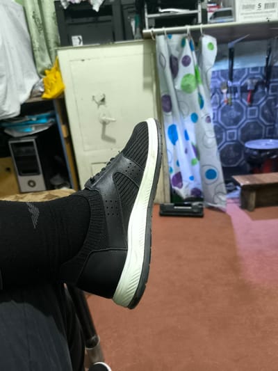 The image shows a person's left foot wearing a black sock and a black sneaker with a white sole, positioned in the foreground and angled towards the right. The background is cluttered and appears to be an indoor setting, possibly a room or storage area, with various objects including what looks like a safe, a shower curtain with circular patterns, shelves with items, and a wooden bench. The overall impression is of a casually organized living or working space. The location of Balkh, Tajikistan is not discernible from the visual information present in the image.