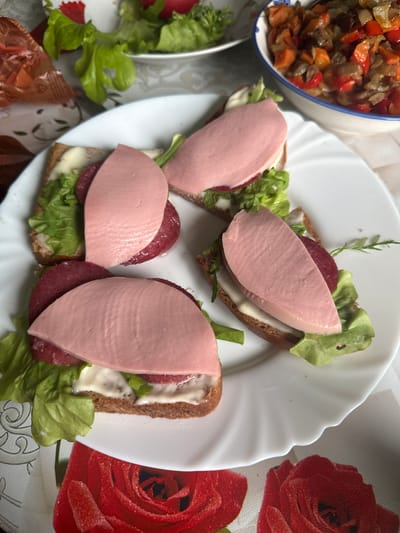 Four open-faced sandwiches are arranged on a white, fluted plate. Each sandwich consists of a slice of dark bread topped with mayonnaise, a lettuce leaf, a slice of salami, and a thick slice of pink, processed meat, possibly bologna. In the background, a bowl of mixed vegetable stew or ratatouille is visible, along with a bowl of lettuce. The scene is set on a tablecloth with a red rose pattern. The image appears to be taken indoors. The prompt mentions the city of Красное, Russia, but there is no visual information in the image to confirm or deny this specific location; the scene is generic and could be anywhere.