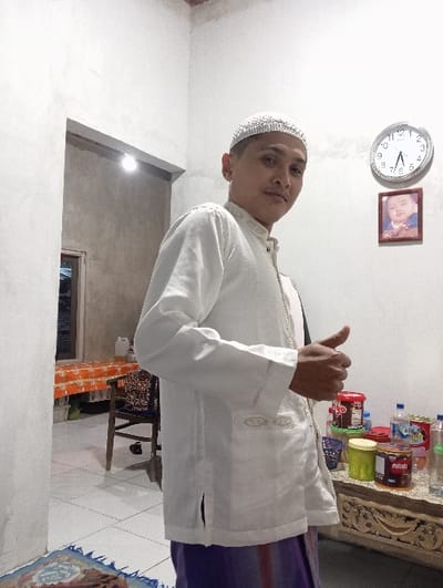 An image taken indoors in Bulukandang, Indonesia, depicts a male subject standing in a room with white walls and a light-colored tiled floor. The subject, positioned slightly left of center and facing the camera, is wearing a white long-sleeved Baju Koko with embroidered patterns, a white peci, and a purple and brown patterned sarong. He is performing a thumbs-up gesture with his right hand.
To the subject's left, an open doorway reveals an adjacent room with unpainted concrete walls. A single bare lightbulb illuminates the doorway. Further into this room, a table draped with an orange and white checkered tablecloth and a wooden chair are visible. A white plastic container rests on the table. A patterned rug is partially visible on the floor in the foreground, bottom left.
To the subject's right, a carved wooden table is laden with various items, including multiple plastic containers (red, green, brown), plastic water bottles, and other packaged goods. Above this table on the white wall, a circular analog clock displays approximately 7:55, and beneath it, a framed picture of a baby is affixed to the wall.