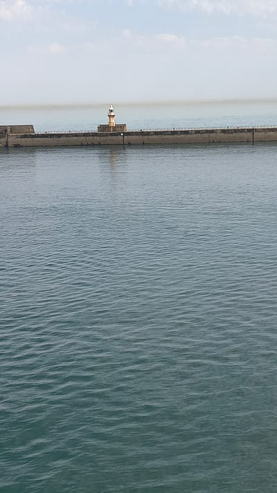 The image shows a wide view of the sea with a breakwater in the distance. On the breakwater, there is a lighthouse. The water is a deep blue-green with visible ripples. The sky is light blue with some faint clouds. The breakwater is made of concrete and extends across the frame. There are some small structures and railings on top of the breakwater. The overall impression is a calm and serene coastal scene.