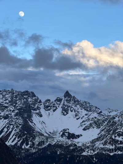 The image depicts a mountain range partially covered in snow. The sky to the left is blue with some clouds, and the full moon is visible. The location appears to be in a mountainous region, possibly near Poschiavo, Switzerland, given the alpine characteristics. There are no people visible in the image. The main subjects are the mountain, sky, clouds, and the moon. The interaction is the interplay of natural elements: mountains meeting the sky, snow on the mountains, clouds partially obscuring the blue sky. The environment is an alpine, high-altitude area.