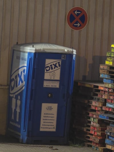 In Zeppelinheim, Germany, a blue portable toilet unit stands against a corrugated metal wall. The unit has the brand "Dixi" printed on it in white, along with images of two figures, one male and one female. There is also a sign on the unit with text in German. To the right of the toilet unit, several stacks of colorful wooden pallets are visible. A round red and blue traffic sign with white arrows is mounted on the wall above and to the right of the portable toilet.