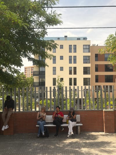 Four individuals are positioned on a tiled surface in front of a brick wall and a metal fence. To the left, a person wearing a black shirt and beige pants sits on the wall, looking down at a phone. To their right, on a white bench, a person with blonde hair, wearing a dark tank top and blue jeans, reads a book. Next to them, another person in a red shirt and black pants holds a phone. Further to the right, a fourth person in a dark shirt and white striped pants sits on the bench, looking at their phone. Behind the individuals, modern apartment buildings with light-colored facades and dark windows are visible, with trees and greenery interspersed. This scene appears to be in Cerdanyola del Vallès, Spain, likely in a residential or public urban area.