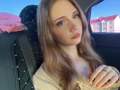 A young woman with long, light brown hair, light makeup, and a light-colored top is sitting in the passenger seat of a car. She is looking at the camera with a neutral expression. The interior of the car is visible, including the black seat with diamond stitching and a seatbelt. Outside the car window, a light pink building with a red roof and blue sky can be seen. This building is located in the city of Novaya Otradovka, Russia. A pink blanket is visible on the back seat. A ring is on the woman's left hand.