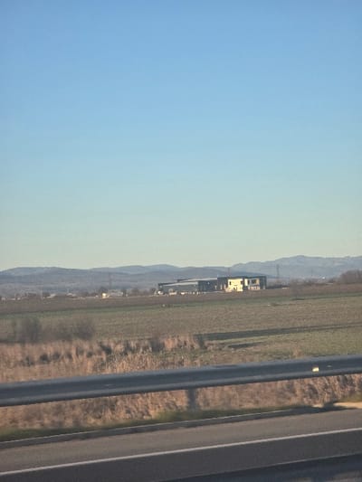 The scene depicts a rural landscape near Diadovo, Bulgaria, observed from a road. In the immediate foreground, a dark grey metal guardrail spans horizontally, positioned above a segment of a paved road with visible white lane markings. Beyond the guardrail, the midground consists of extensive fields displaying brown and green vegetation, interspersed with patches of dry, reedy growth and sparse, bare trees or bushes. In the distant background, a prominent industrial-style building complex is visible within these fields, composed of a large, dark grey structure with an attached lighter-colored section. A range of low, undulating mountains forms the furthest horizon. The upper two-thirds of the image are occupied by a clear, gradient pale blue sky, indicative of daylight. No individuals or discernible activities are present.