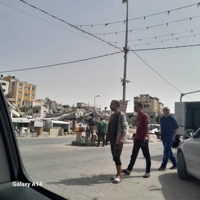 The image depicts a street in Nuseirat, Palestinian Territories, with visible damage to buildings. Three men stand in the foreground. Behind them, there is debris and destroyed structures. A car is partially visible in the foreground. Overhead, there are electrical lines and light fixtures.