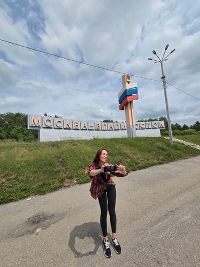 A woman stands on an asphalt road, holding a smartphone in landscape orientation to take a photo. She is wearing a red and black plaid shirt, black leggings, and black and white athletic shoes. She is positioned in front of a large sign that reads "МОСКВА-ВЛАДИВОСТОК" in large, orange, raised letters against a light gray background. To the right of the sign is a tall, light-colored monument with horizontal stripes in white, blue, and red, resembling the Russian flag. A street lamp with multiple bulbs stands near the monument. The background features a grassy slope with scattered yellow wildflowers, trees, and a cloudy sky. The location is identified as the city of Nikolaevka, Russia.