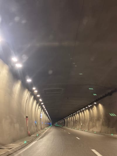 The image shows the interior of a tunnel, likely a road tunnel, as evidenced by the asphalt road surface and lane markings. The tunnel is well-lit by rows of lights along the ceiling and walls. Some lights on the ceiling appear to be bright white, while others further down the tunnel emit a greenish hue. Along the base of the tunnel walls, there are small green lights, possibly delineating the edge of the road or a walkway. The walls themselves are a rough, textured concrete, and the ceiling is also concrete. The perspective suggests the photo was taken from a vehicle moving through the tunnel. In the distance, the tunnel curves slightly to the right. There are no people or other vehicles visible in the frame. The overall impression is of a functional and modern tunnel infrastructure. The specific mention of Surami, Georgia, suggests this is a known tunnel in that location, although the image itself does not contain any explicit geographical markers.