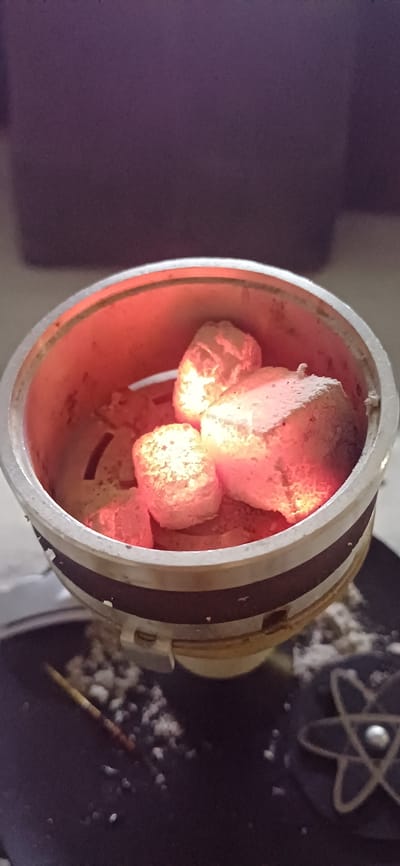 The image shows a close-up of a hookah bowl filled with glowing red coals. The coals are cylindrical and covered in white ash. They are emitting a strong red glow, indicating they are very hot. The bowl itself is metallic with dark stripes and a white plastic piece attached to the side. Ashes are scattered around the bowl, and a wooden stick or skewer is visible on the left. In the background, a dark purple surface is visible, and on the right side, a dark object with a star-like symbol can be seen. The scene appears to be indoors, with artificial lighting contributing to the glow of the coals. There is no visible indication of Zuevka, Russia, or any specific urban context.