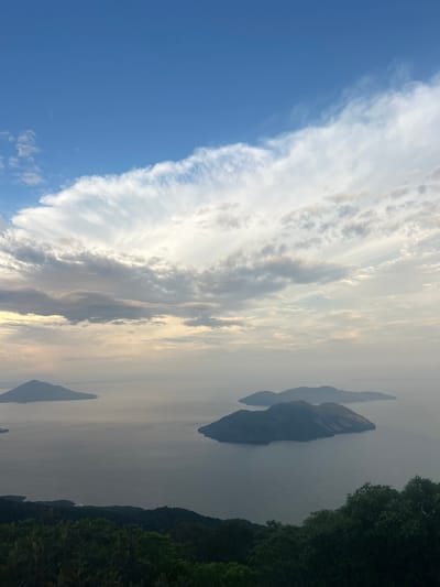 This is a high-angle, wide shot of a seascape with multiple islands under a cloudy sky. The foreground consists of dark green foliage, suggesting a hillside or elevated viewpoint. The middle ground is dominated by a large body of water, appearing calm and reflecting the light from the sky. Several islands of varying sizes are scattered across the water, their dark shapes contrasting with the muted tones of the sea and sky. The background is a vast expanse of sky, filled with a mix of bright white and gray clouds, with patches of clear blue visible. The lighting suggests it is either early morning or late afternoon. The image captures a natural landscape, likely a coastal or island region. Without specific landmarks or identifying features in the image, it is impossible to determine the exact location within El Cacao, El Salvador, or any other city.