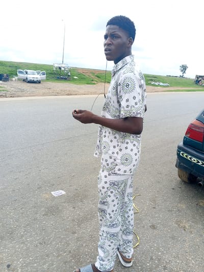The image shows a young Black male standing on an asphalt road in Pasere, Nigeria. He is wearing a white, patterned shirt and matching pants, and grey sandals. The man is looking to his left, with a slightly concerned expression. He is holding a small black object in his right hand, possibly earbuds. In the background, there is a grassy embankment with a few scattered objects, including a car with its doors open, a makeshift shelter with a light pole, and some tires. To the right of the man, the rear of a dark-colored car is visible. The sky is overcast, suggesting a cloudy day. The scene appears to be outdoors, possibly near a roadside car wash or repair area. A small piece of white paper with visible text is on the road in front of the man. The text on the paper is partially obscured but appears to read "ALLOY".