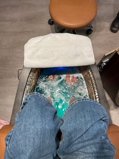 From an overhead perspective, the image shows a pair of feet immersed in a foot spa filled with bubbling, blue-tinted water. The individual is wearing blue denim jeans. A rolled white towel is placed on the edge of the foot spa. The spa rests on a dark grey surface. To the left is a round, brown padded stool with a chrome base. A black leather boot is visible in the upper left corner. The specific location within Kelowna, Canada, is indeterminable from the image.