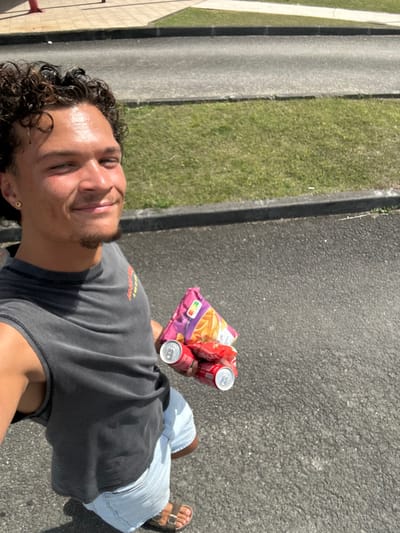 A young male, approximately 20-30 years old, with dark curly hair and a short goatee, is captured in a sunlit selfie against an outdoor urban backdrop. He has a light complexion with a sheen of moisture on his forehead, and he is smiling directly at the camera. He wears a dark grey sleeveless t-shirt, light blue denim shorts, and brown slide sandals. A gold stud earring is visible in his left earlobe.
In his right hand, he holds a purple bag of potato chips, distinguished by yellow and red graphics and a "Nutri-score A" label, along with three red soda cans, likely Coca-Cola, stacked and held with their silver tops visible.
The subject stands on dark asphalt pavement, consistent with a street or parking lot. To his right, a dark grey curb separates the asphalt from a strip of green grass, which is bordered by another dark grey curb. Beyond this, a lighter grey paved surface leads to what appears to be a light-colored building foundation or wall in the upper left background. A red fire hydrant or similar utility fixture is partially visible in the upper left corner, behind the subject's head. The environment is consistent with a generic urban or suburban area within Le Moule, France, though a precise street location is not discernible from the image.