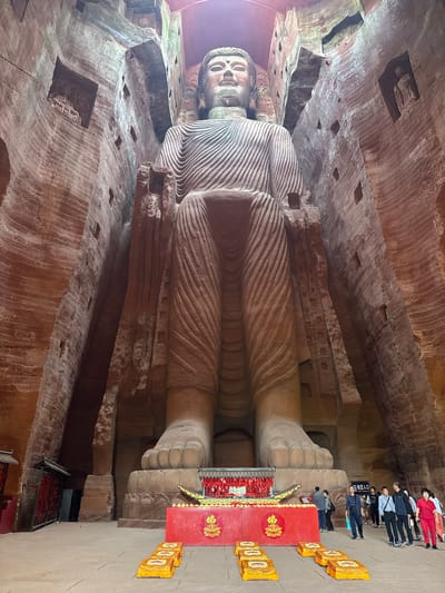 The image shows the Leshan Giant Buddha in Leshan, Shizhong, China. It is a massive stone carving of a seated Maitreya Buddha, carved out of a cliff face, with people standing near its foot, offering candles, and admiring the view. The surrounding rock formations and carved details are visible.